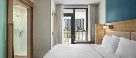 Habitación de hotel con una cama grande, cabina de ducha de vidrio, vista de edificios por la ventana y un cabecero de madera.