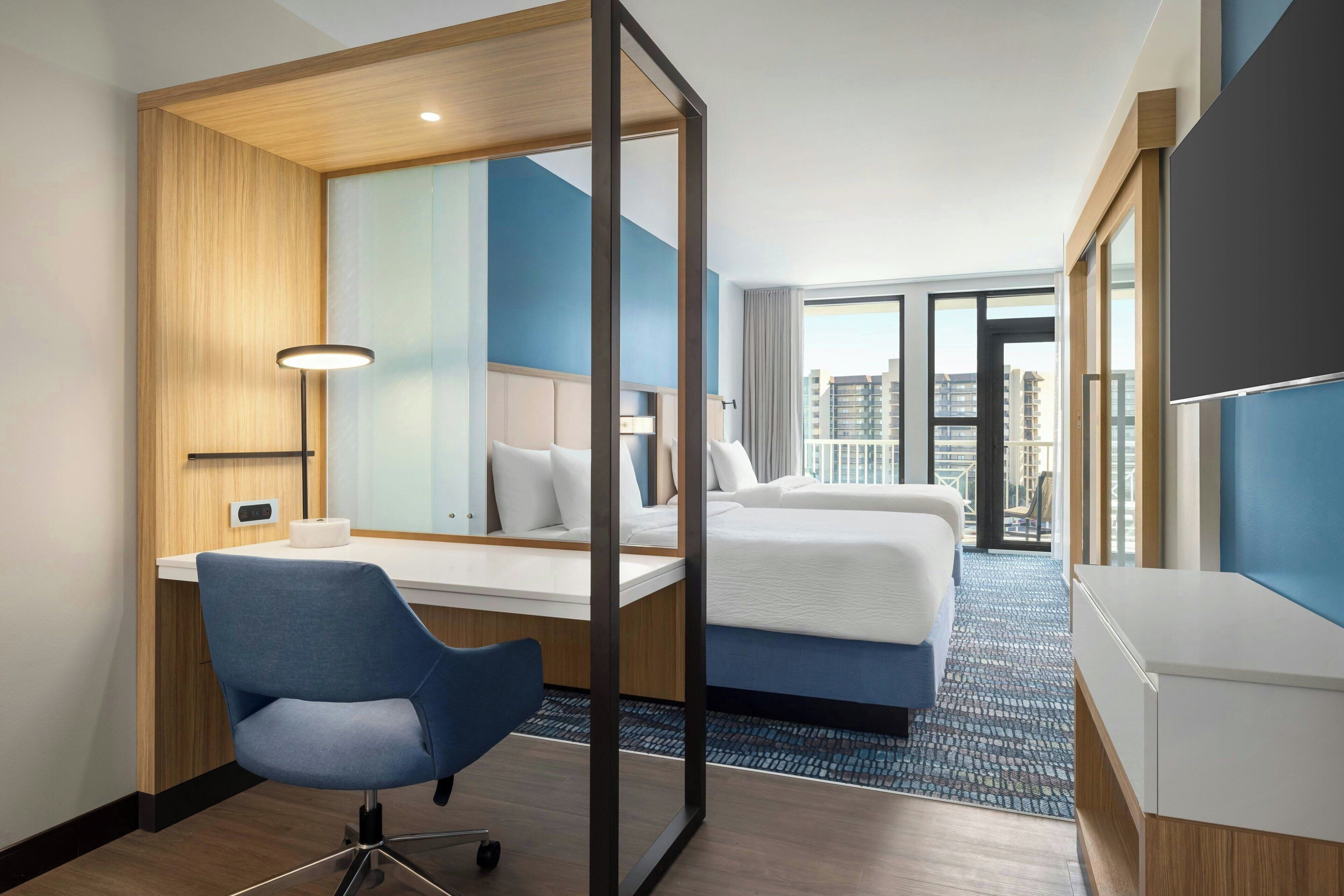 Une chambre d’hôtel équipée d’un bureau, d’une chaise, d’un lit et de grandes fenêtres donnant sur la ville.