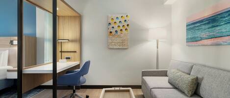 A modern hotel room with a desk, a grey sofa, a small table, a chair, a painting, and a lamp.