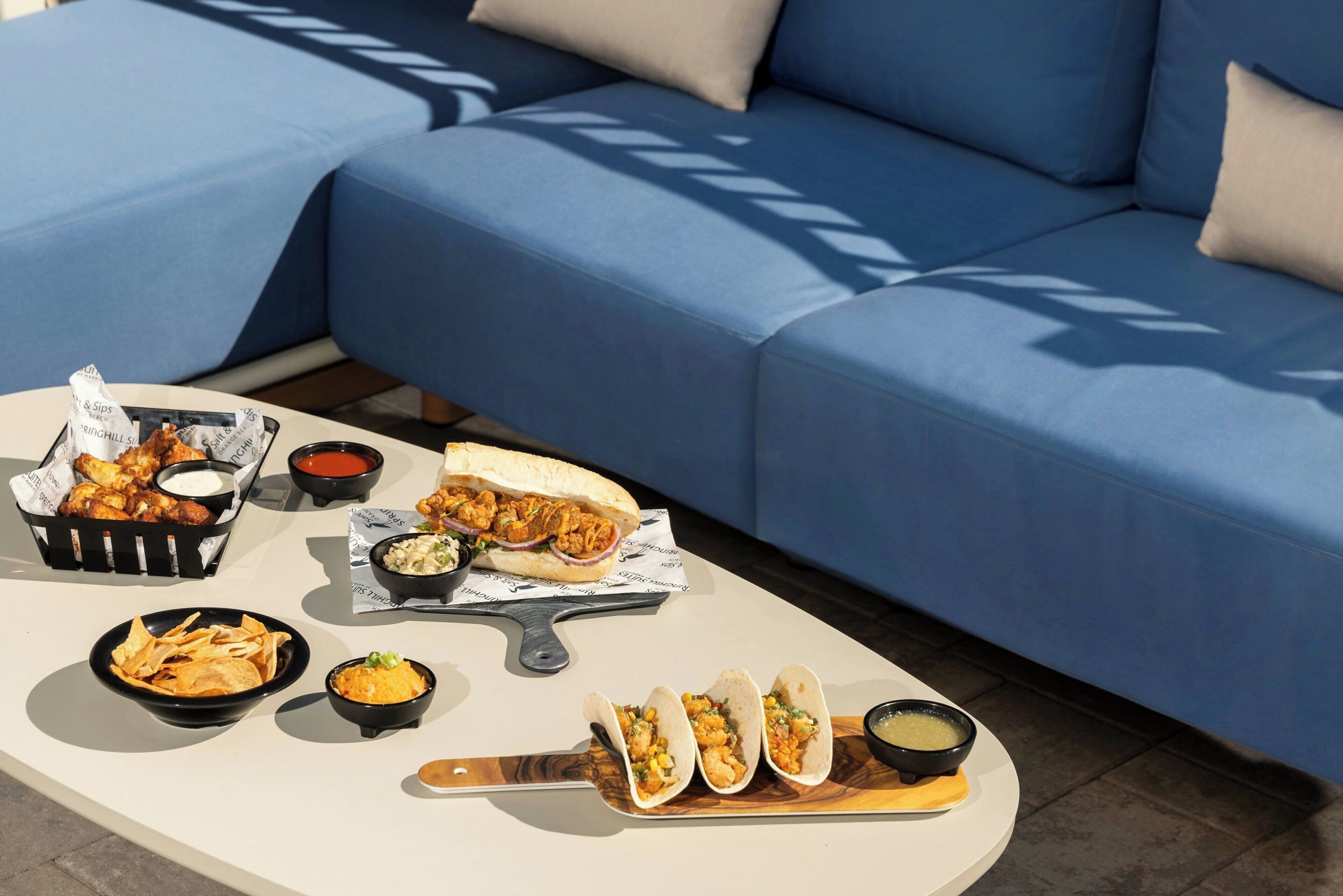A dining table with various food items including tacos, nachos, and a sandwich, set on a patio with a blue sofa in the background.