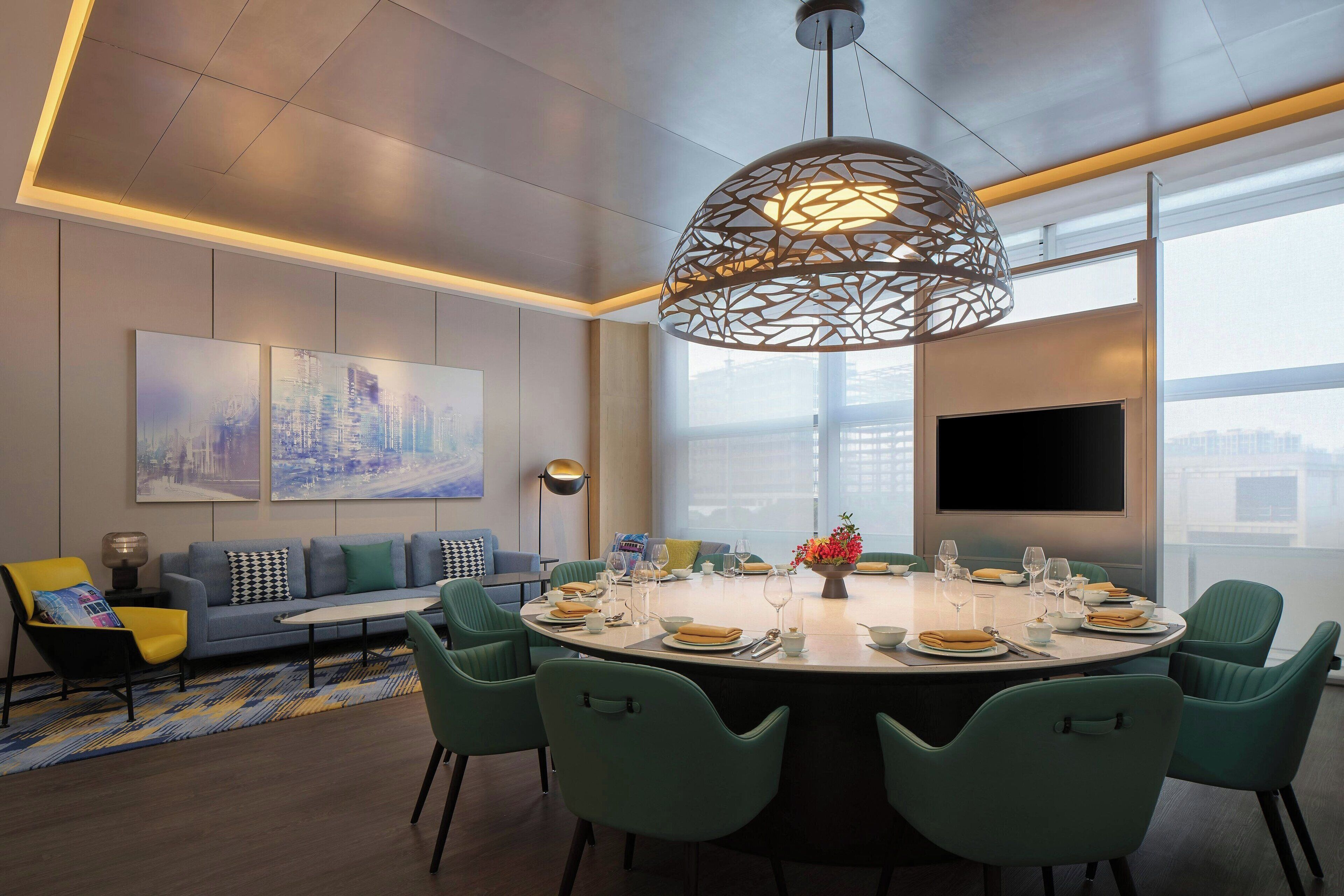 A modern dining area with a round table set for six, surrounded by teal chairs, a sofa with patterned cushions, and a cityscape painting on the wall.