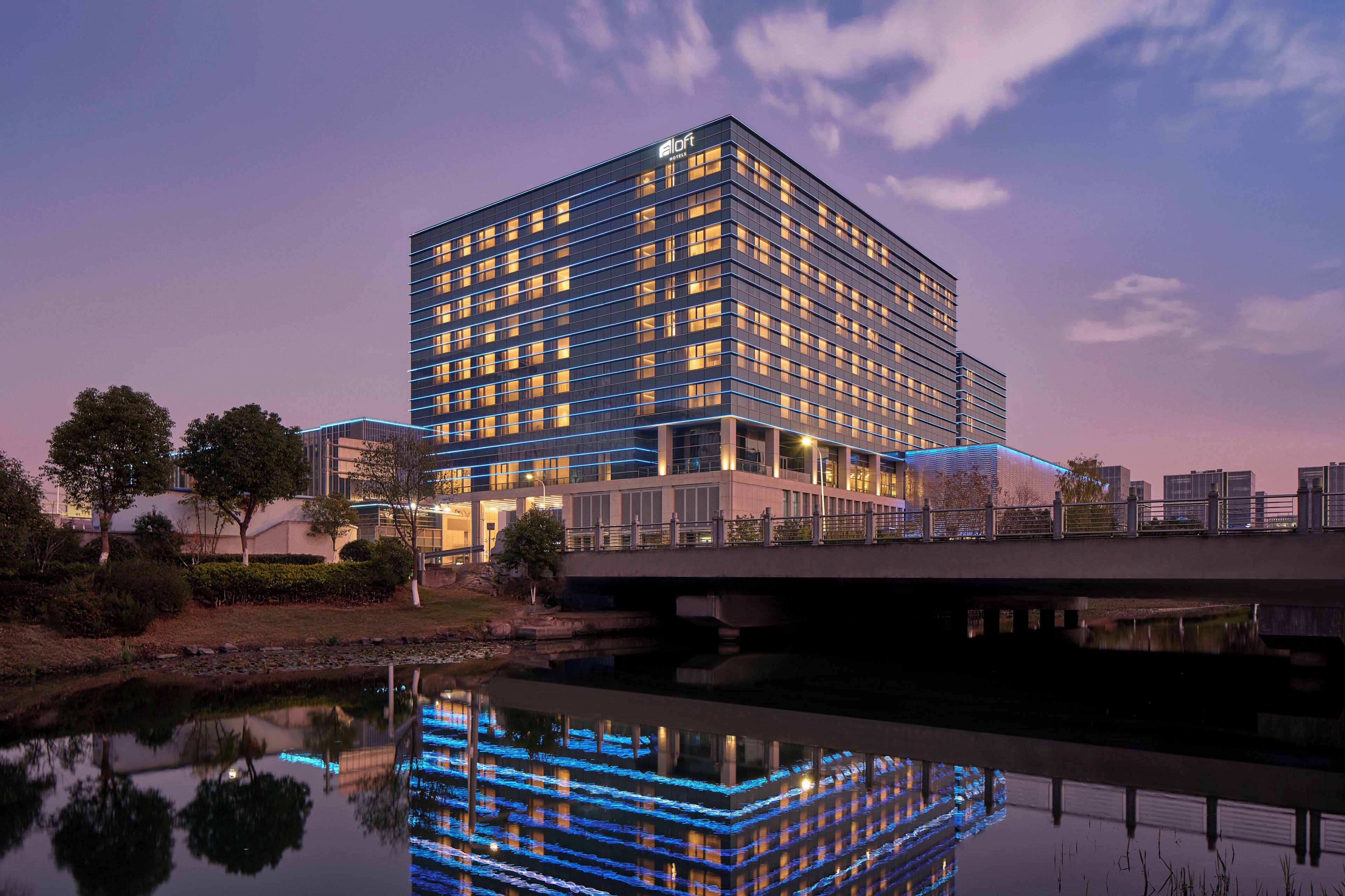 Un hotel moderno con una piscina reflectante, un puente y un horizonte urbano de fondo.