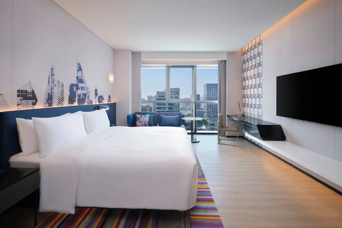 A modern hotel room with a large bed, a TV, a city view, and a patterned wall.