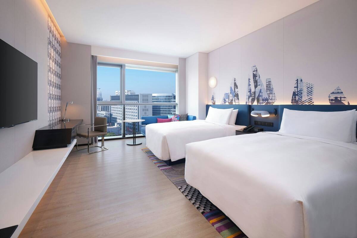 A hotel room with two beds, a desk, a chair, a TV, and a large window with a city view.