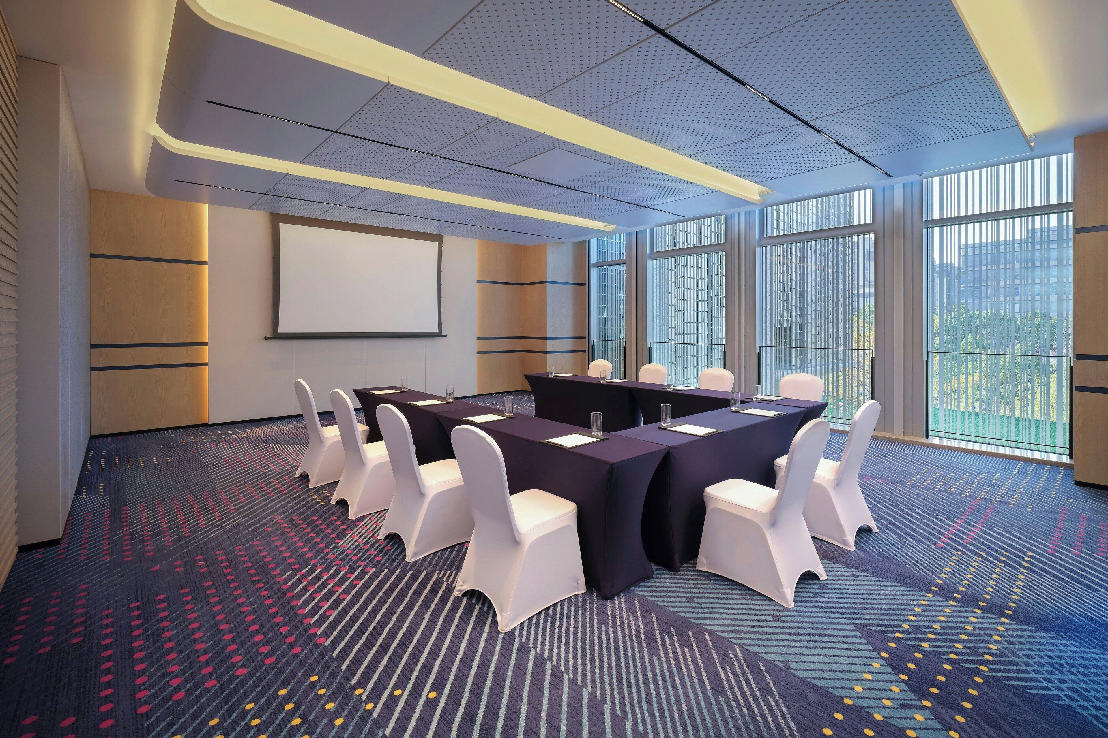 A conference room with a large table, white chairs, and a projector screen.