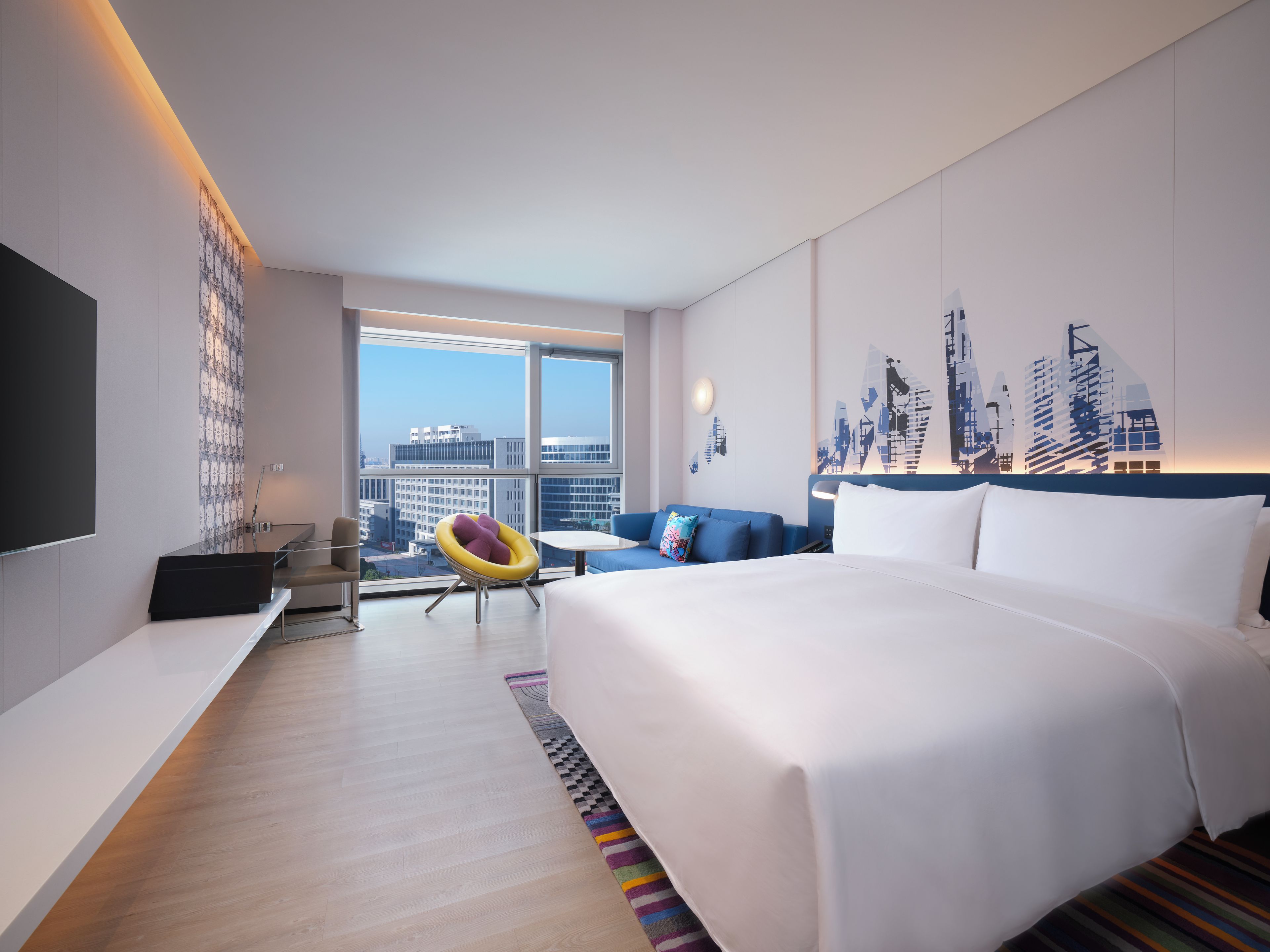 A hotel room with a large bed, a desk, a chair, and a view of the cityscape.