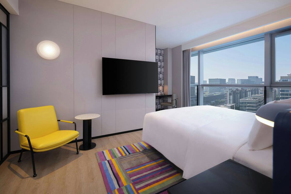 A hotel room with a bed, a yellow chair, a small table, a TV mounted on the wall, and a city view through large windows.