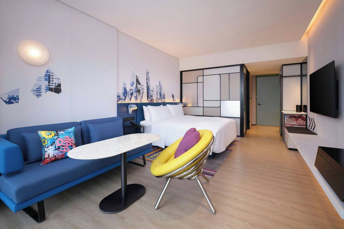 A modern hotel room with a blue sofa, a small round table, a unique yellow and purple chair, a bed with white bedding, a TV mounted on the wall, and a cityscape mural on the wall.