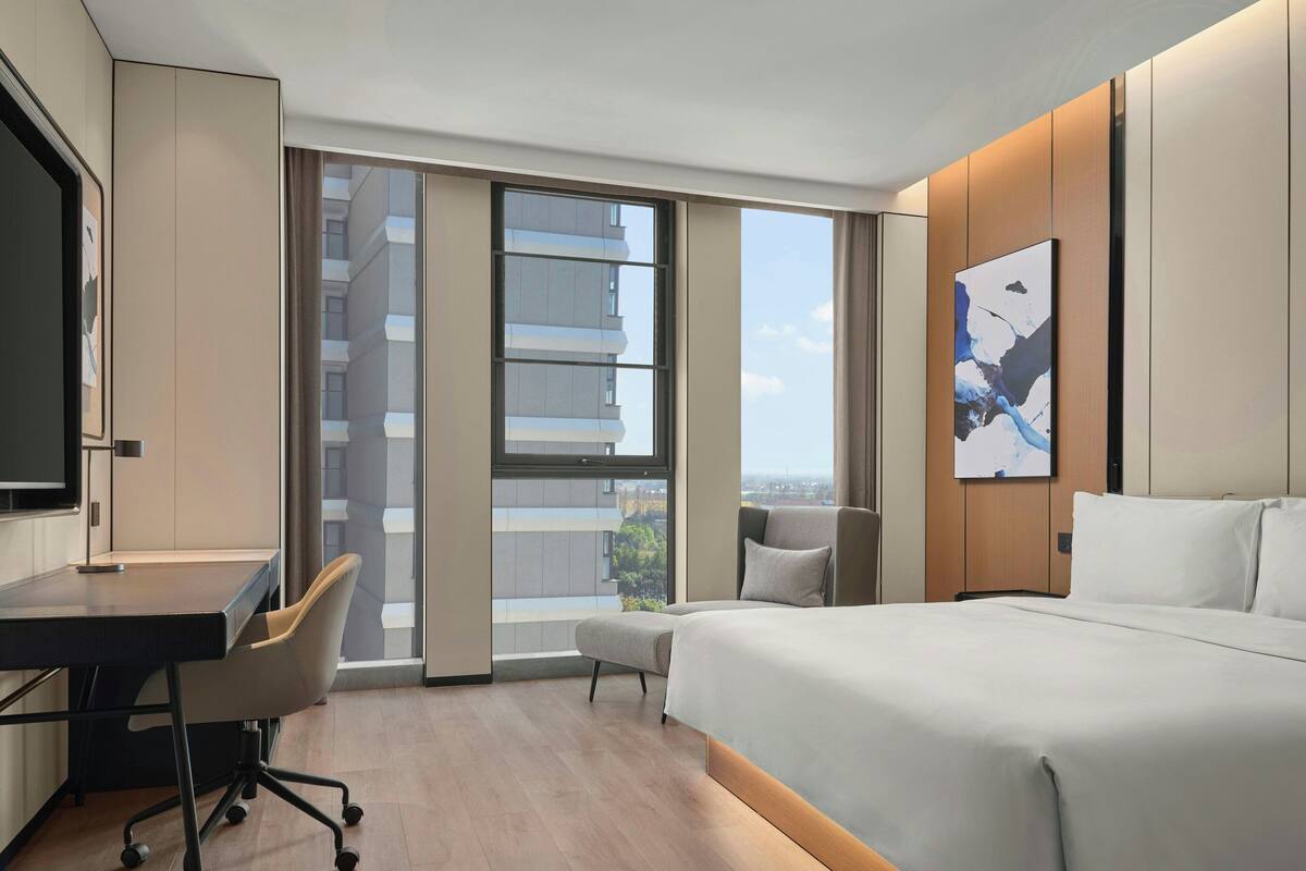 Una habitación de hotel moderna con una cama grande, un escritorio y una silla. Hay un ventanal con vistas a edificios.
