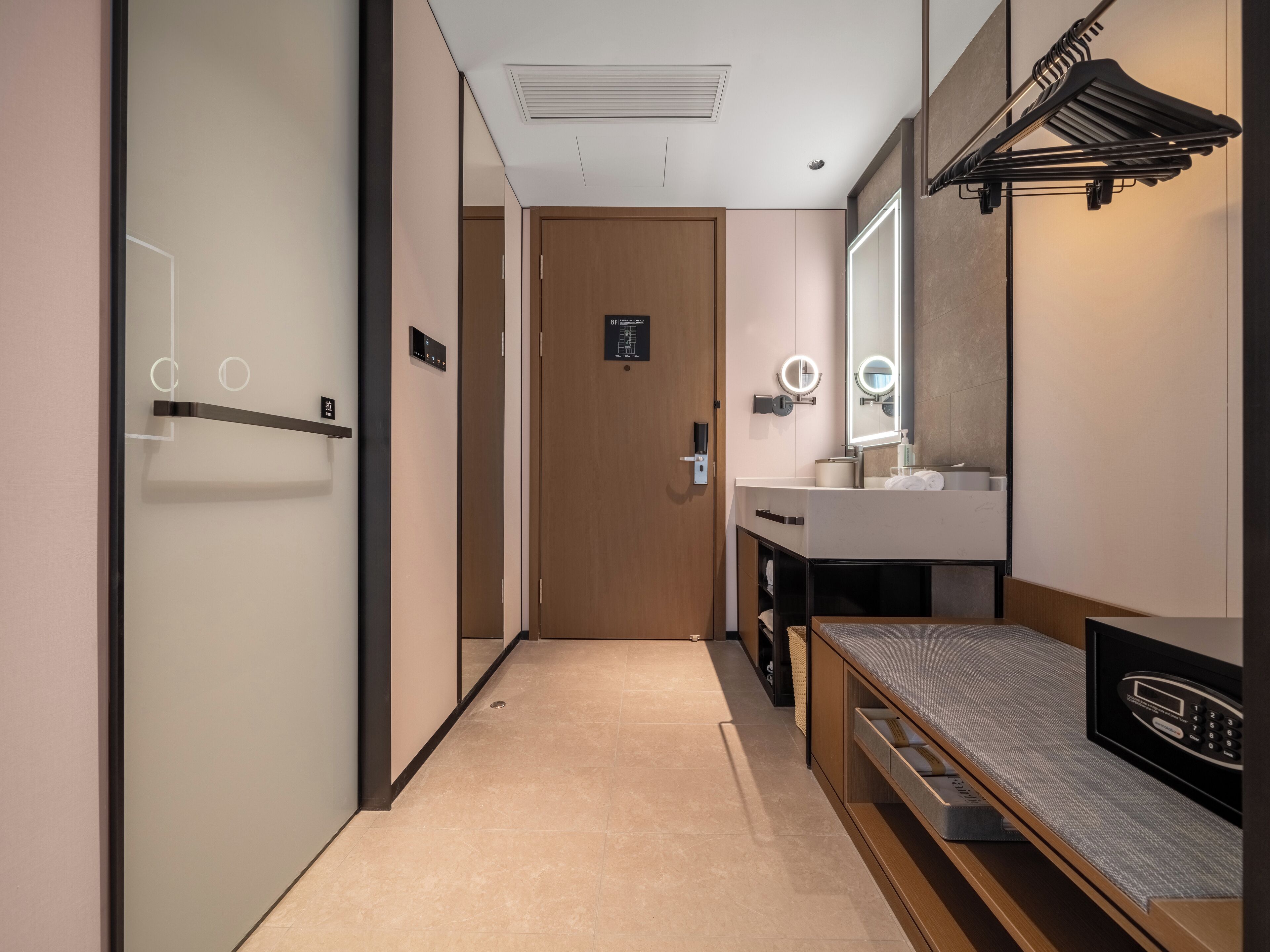 A modern hotel room with a walk-in closet, a vanity with a mirror, and a bed with a control panel.