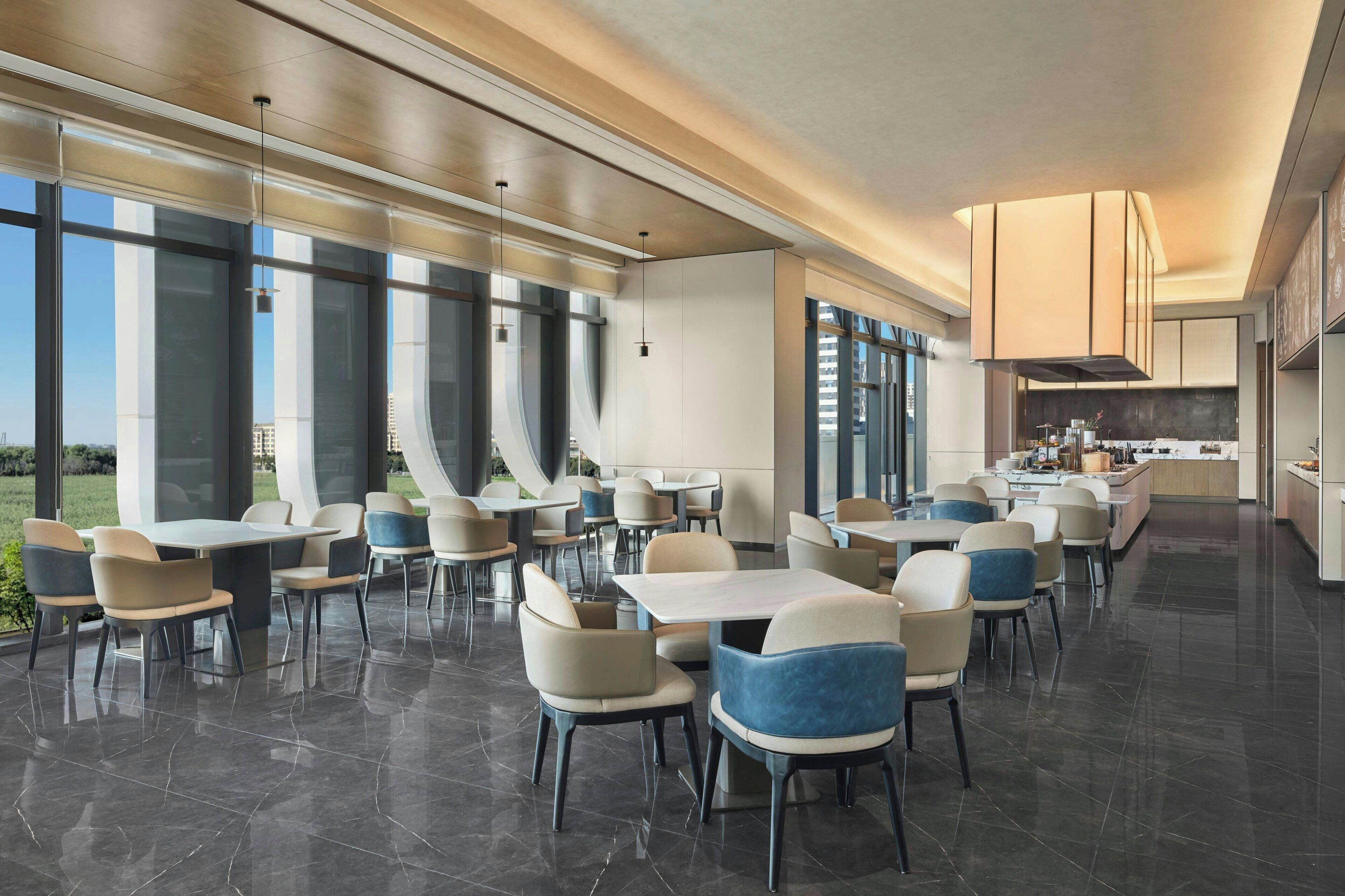 Un espace de restauration moderne, avec des tables et des chaises, de grandes fenêtres et une cuisine en arrière-plan.
