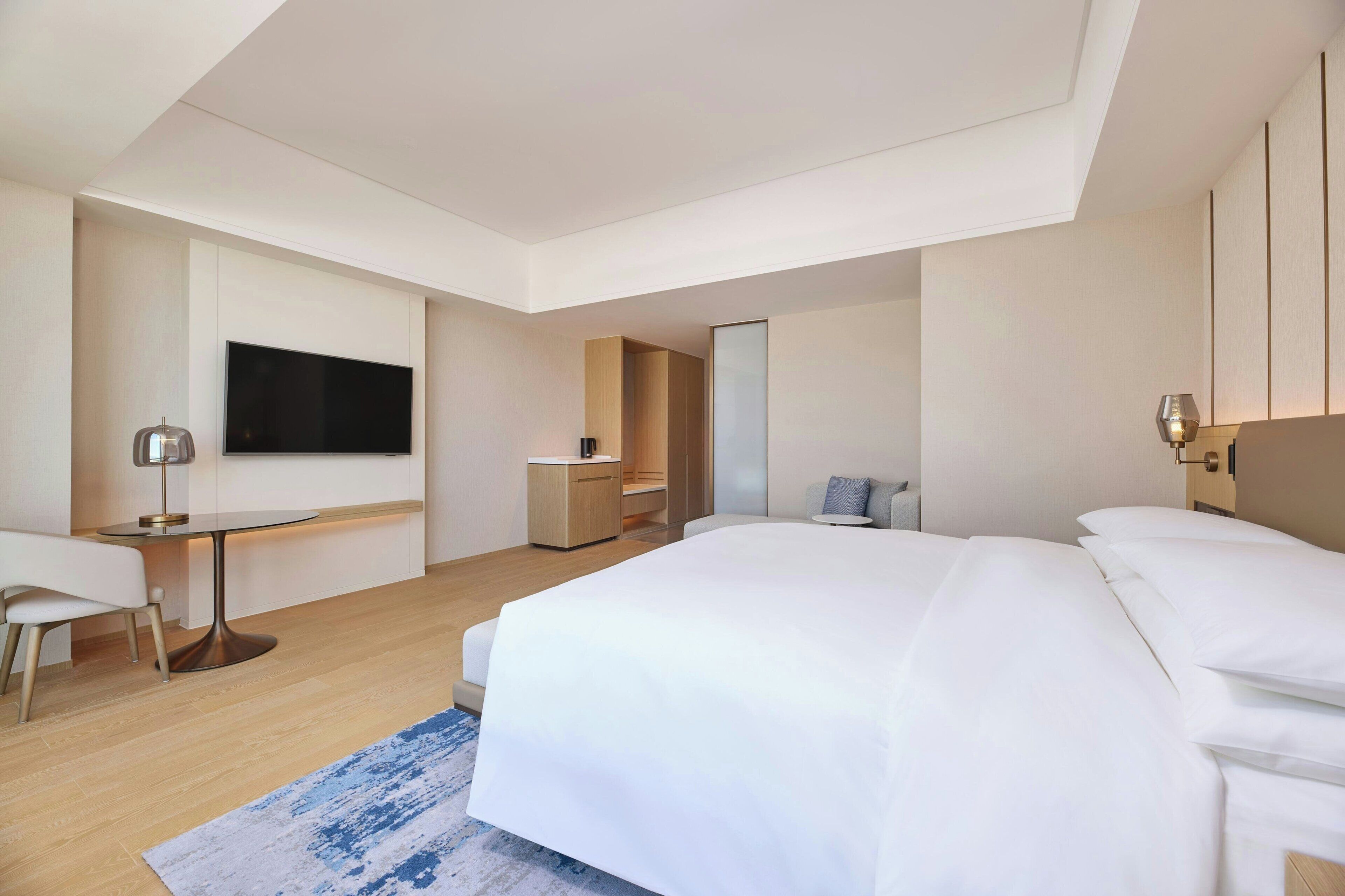 A modern hotel room with a large bed, a desk, a chair, a TV, and a small seating area.