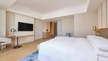 A modern hotel room with a large bed, a desk, a chair, a TV, and a small seating area.