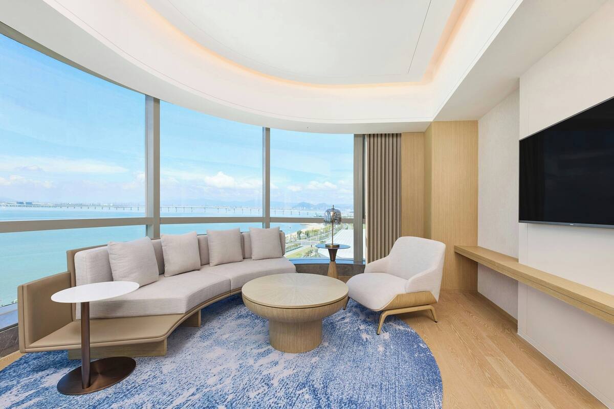 Une chambre d’hôtel moderne avec un plafond incurvé, de grandes fenêtres donnant sur un pont et l’eau, un canapé, un fauteuil, une table basse et un téléviseur à écran plat.
