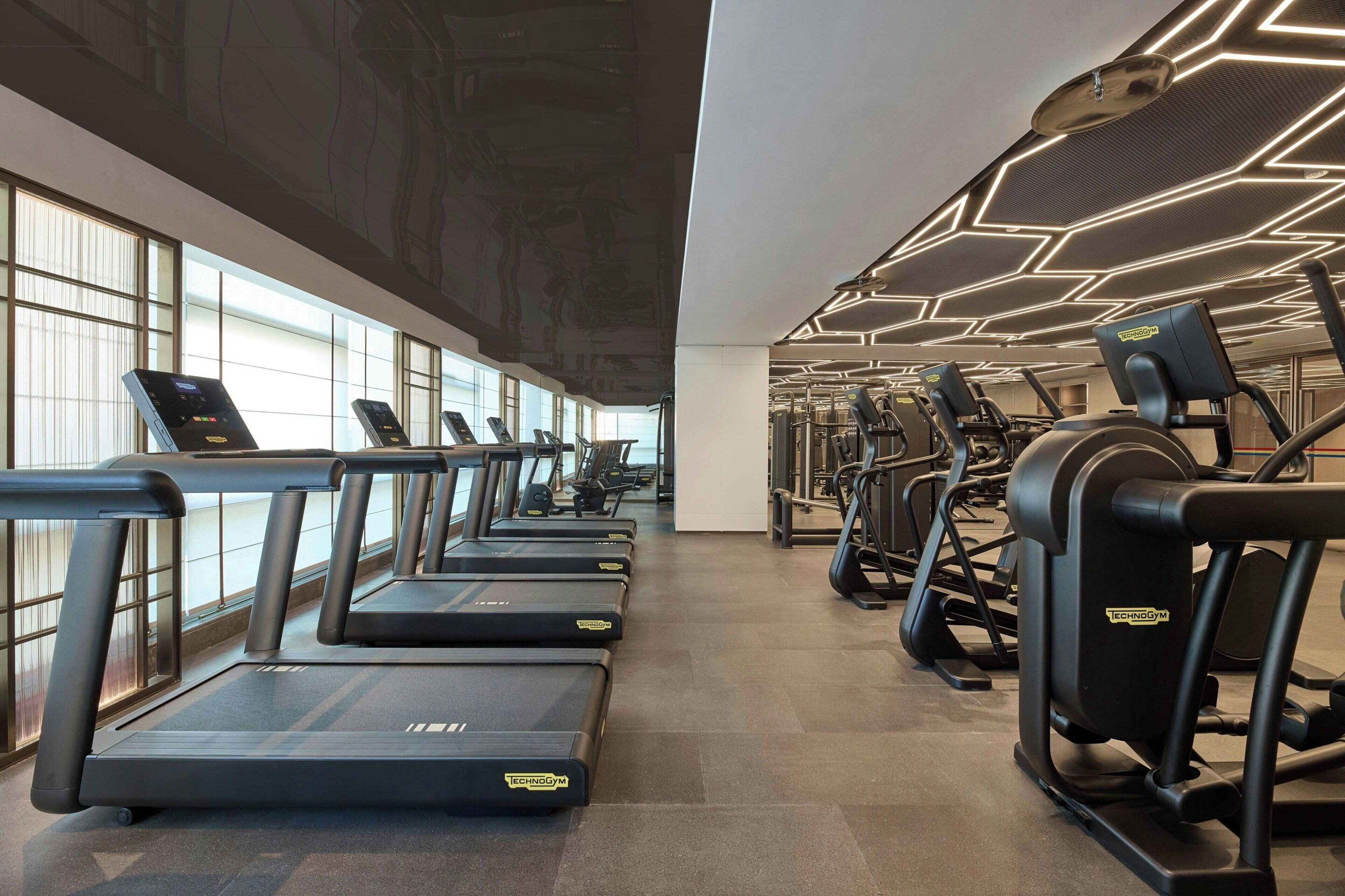 Een sportschool met loopbanden en fitnessapparatuur, grote ramen en een geometrisch plafondontwerp.