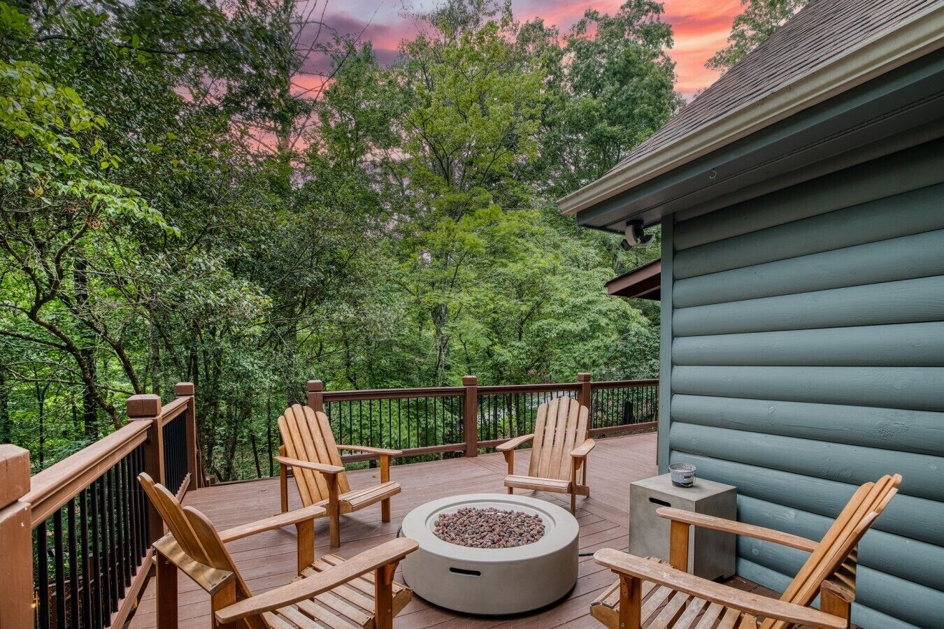 A wooden deck with Adirondack chairs and a fire pit, surrounded by lush greenery and a house with a green exterior.