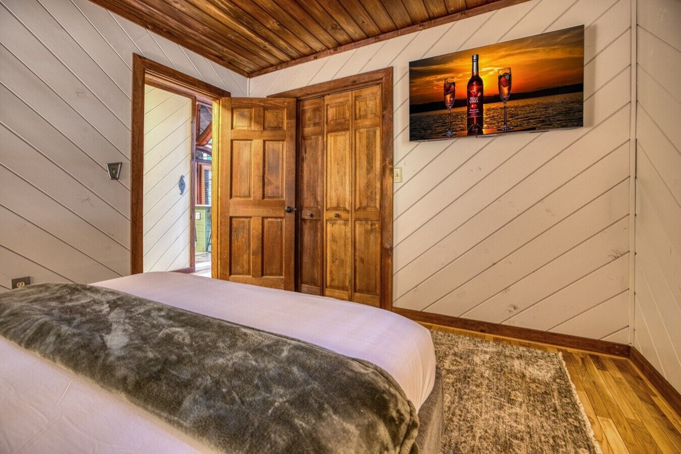 A bedroom with a bed, wooden doors, a painting of wine glasses, and a view of the outdoors.