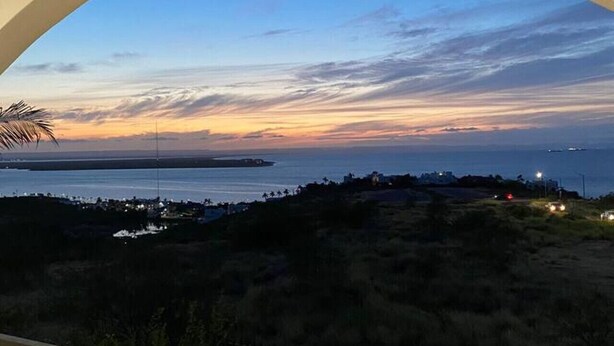 A balcony with a view of a sunset over a coastal landscape, including a body of water and distant land.