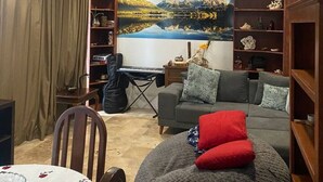 A living room with a grey sofa, a red cushion, a wooden bookshelf, a table with a floral tablecloth, and a large landscape painting on the wall.