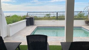 A covered outdoor area with a pool, two lounge chairs, and a view of the sea.