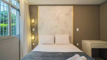 A hotel room with a bed, a bedside table, a window with curtains, and a wall-mounted light fixture.