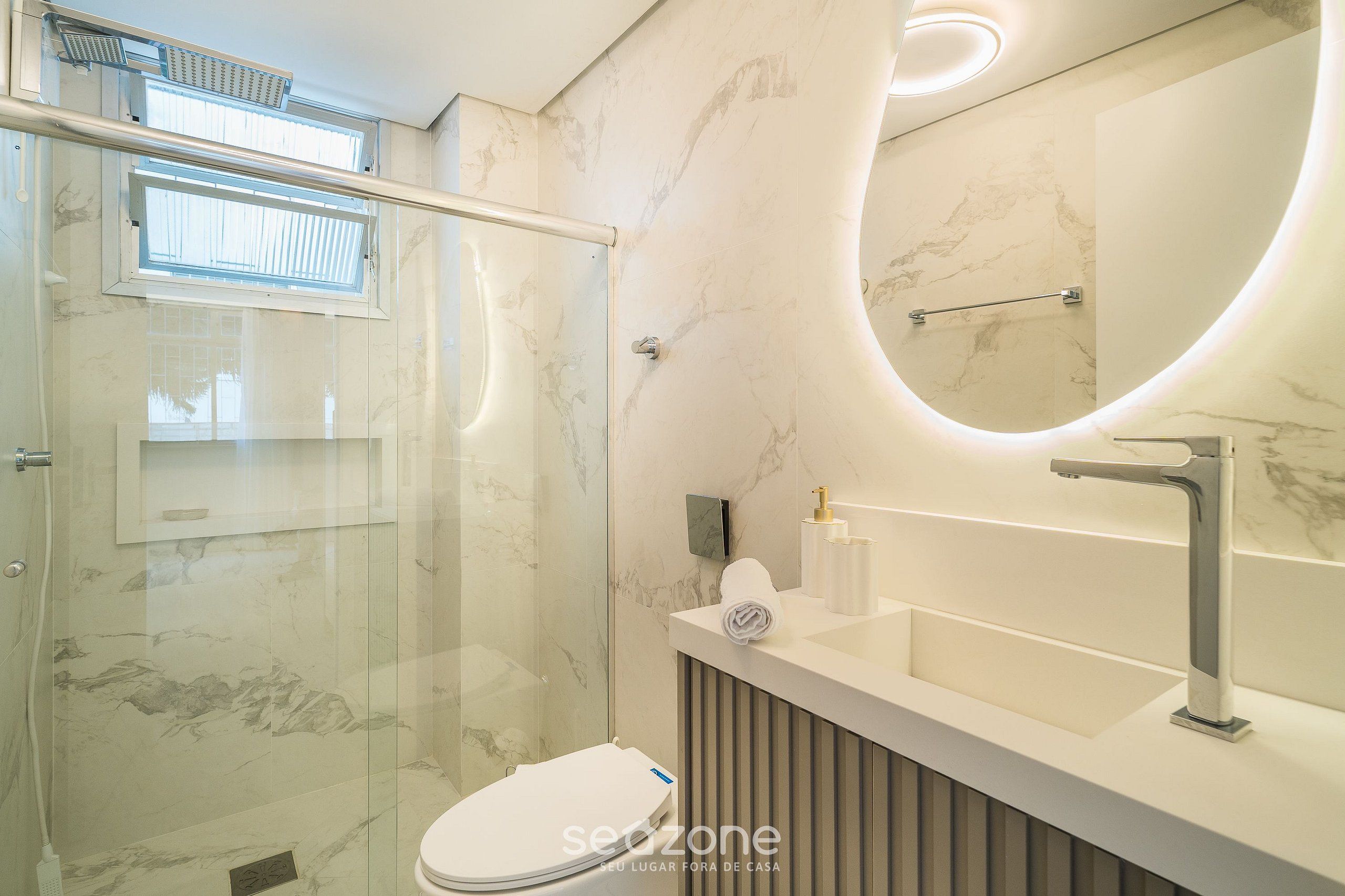 A modern bathroom with a glass shower enclosure, a white sink with a marble countertop, and a large mirror with a curved frame.