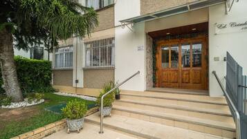 A building entrance with a wooden door, steps, a metal handrail, and a small garden area with potted plants.