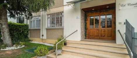 A building entrance with a wooden door, steps, a metal handrail, and a small garden area with potted plants.