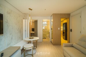 A modern living room with a sofa, a round table, and a bench. There is a TV mounted on the wall, and a wooden panel separates the living area from the kitchen. - Full apt Near Ufsc in Florianopolis Cec0612 (Florianópolis)
