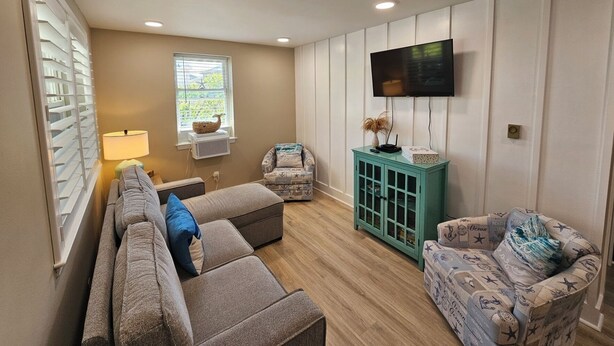 Un salon comprenant un canapé, un fauteuil et un meuble de couleur turquoise. Un téléviseur est fixé au mur, et une fenêtre avec des stores est visible.