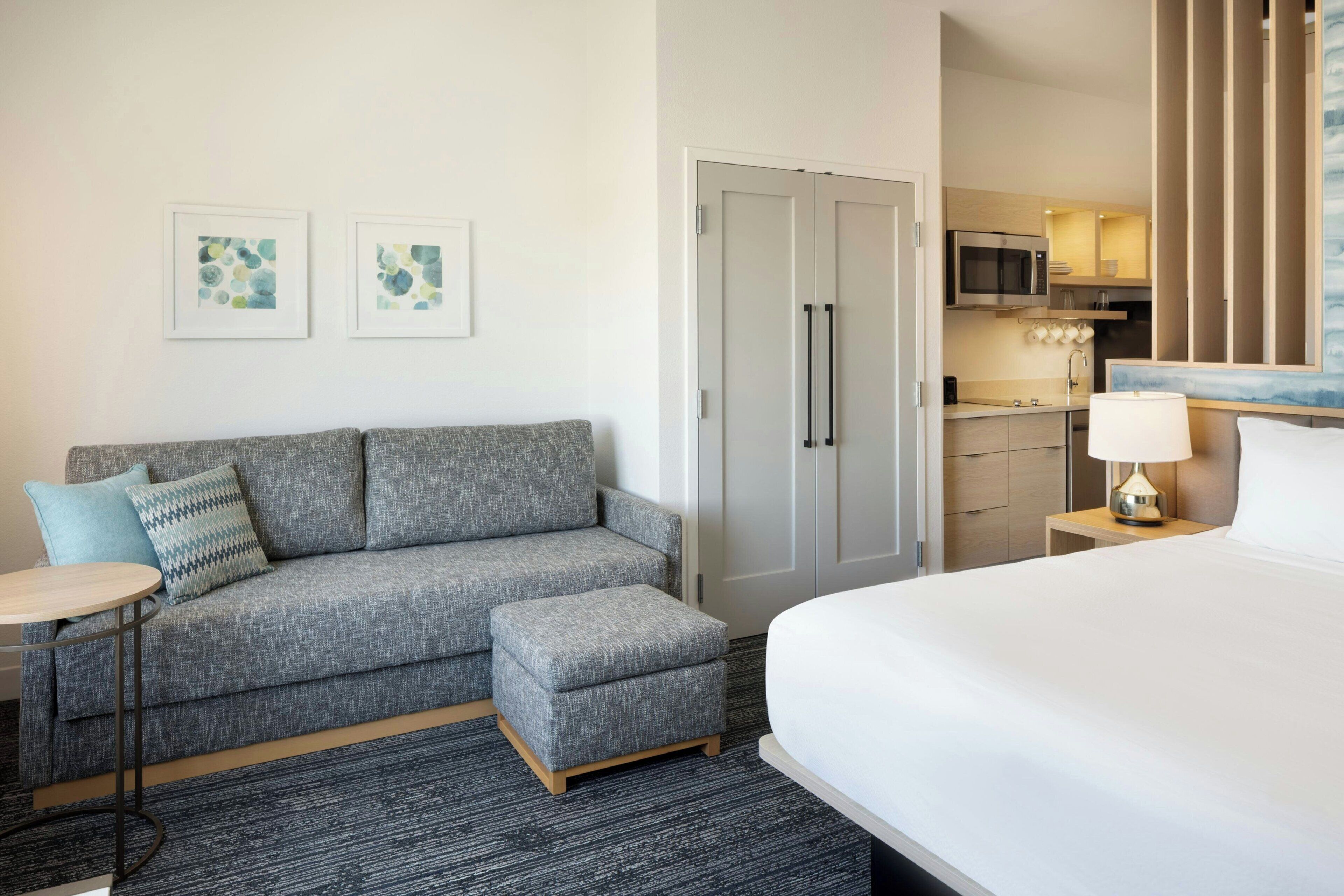 A hotel room with a grey sofa, a small ottoman, a bed, a nightstand with a lamp, a microwave, and a kitchenette.