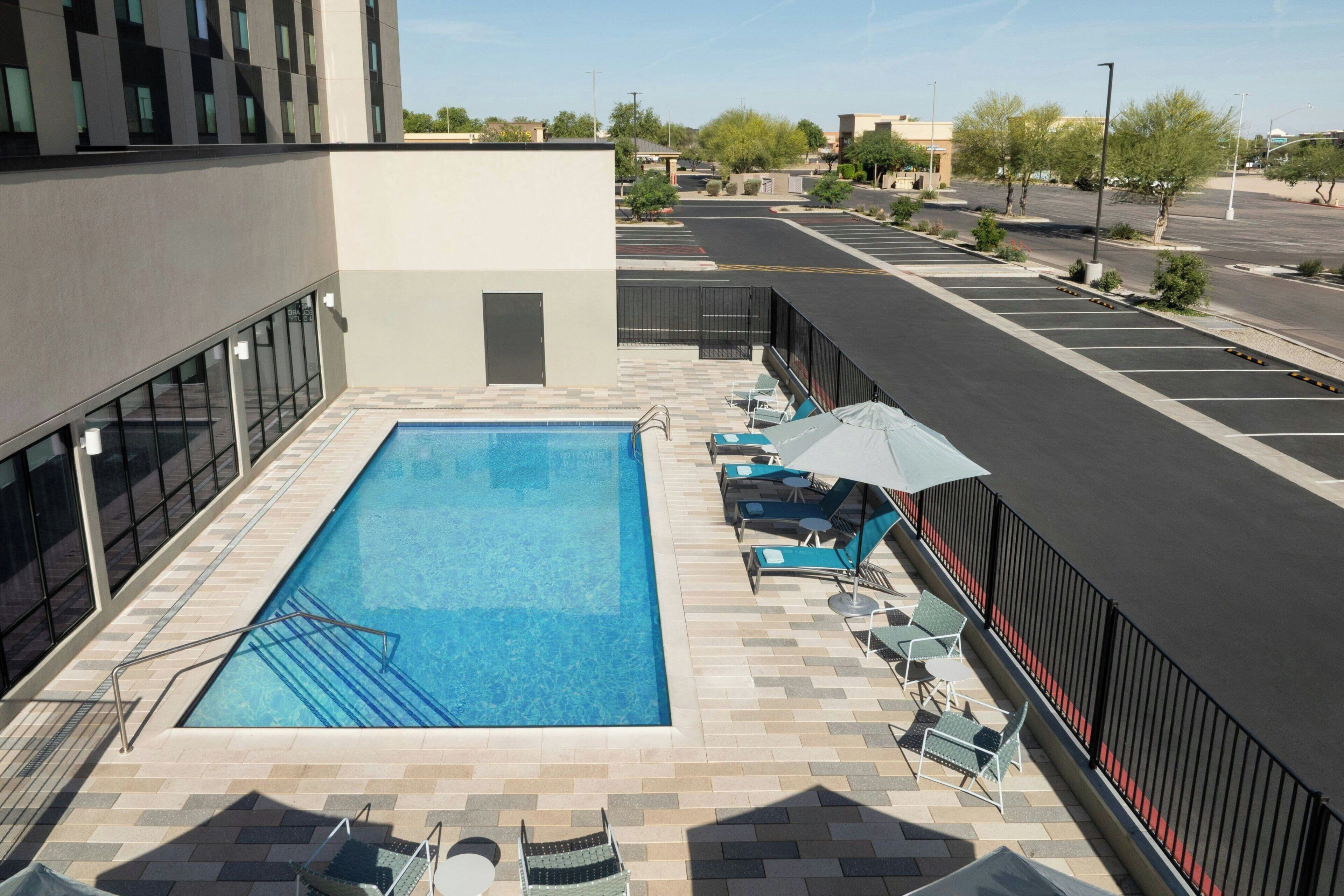 A rooftop swimming pool with lounge chairs and umbrellas, surrounded by a parking area and buildings.