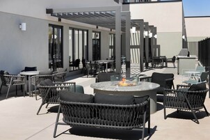 Un patio al aire libre con mobiliario de mimbre, una chimenea central y fachada de edificio moderna.