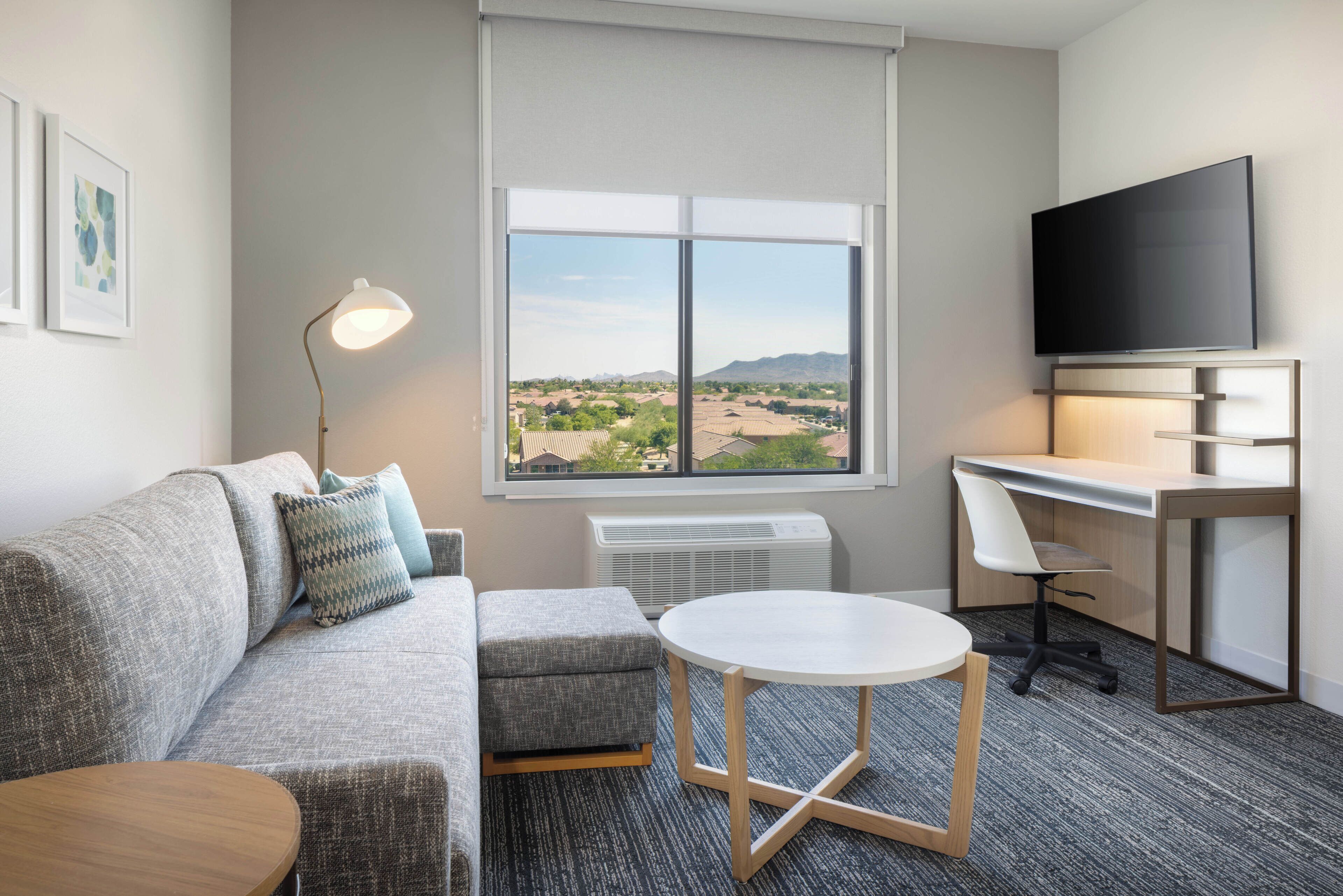 A hotel room with a gray sofa, a small round table, a desk with a chair, a flat-screen TV, and a large window with a view of a residential area.