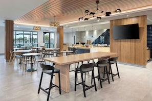 A modern dining area with a long table and multiple chairs, a kitchen area with a flat-screen TV, and a view through large windows. - Towneplace Suites By Marriott Casa Grande (Casa Grande)