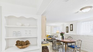 Ein helles, weißes Interieur mit Essbereich, der einen Holztisch und -stühle, eine weiße Schrank mit Regalen und ein Fenster mit Vorhängen umfasst.