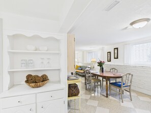 Ein helles, weißes Interieur mit Essbereich, der einen Holztisch und -stühle, eine weiße Schrank mit Regalen und ein Fenster mit Vorhängen umfasst.