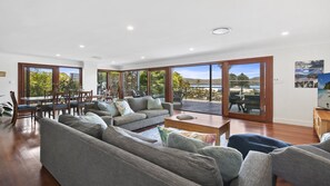A spacious living area with a large sofa set, a coffee table, and a dining table with chairs. There is a sliding glass door leading to an outdoor deck with a view of a lake and trees. - Poolside at Wagstaffe (WAGSTAFFE)