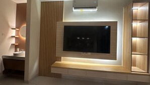 A modern living room with a flat-screen TV mounted on a wooden wall, a built-in shelf below, and a ceiling-mounted air conditioner. - Luxury central Denpasar home with pool and clubhouse for an easy family stay. (Denpasar Barat)