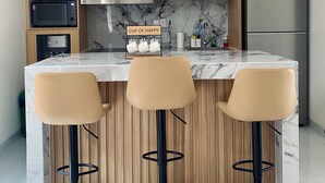 A modern kitchen with wooden cabinets, a marble countertop, a stainless steel refrigerator, and three bar stools. - Luxury central Denpasar home with pool and clubhouse for an easy family stay. (Denpasar Barat)