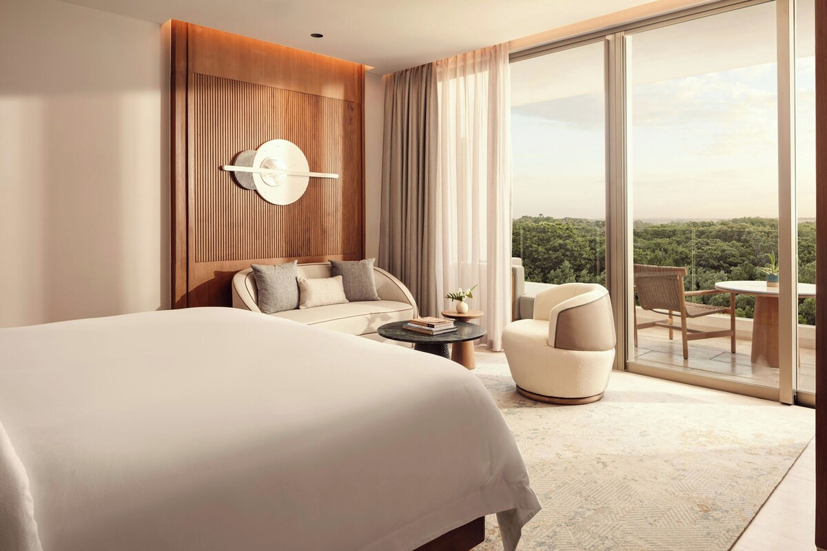 A modern hotel room with a large bed, a seating area with a sofa and armchairs, a small table, and a balcony with a view of the greenery.