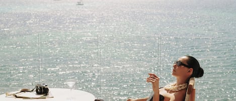 A person seated at a round table with a camera and a drink, overlooking a body of water with boats.