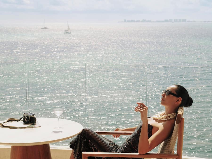 Uma pessoa sentada a uma mesa redonda, com uma câmera e uma bebida, com vista para um corpo d'água e barcos.