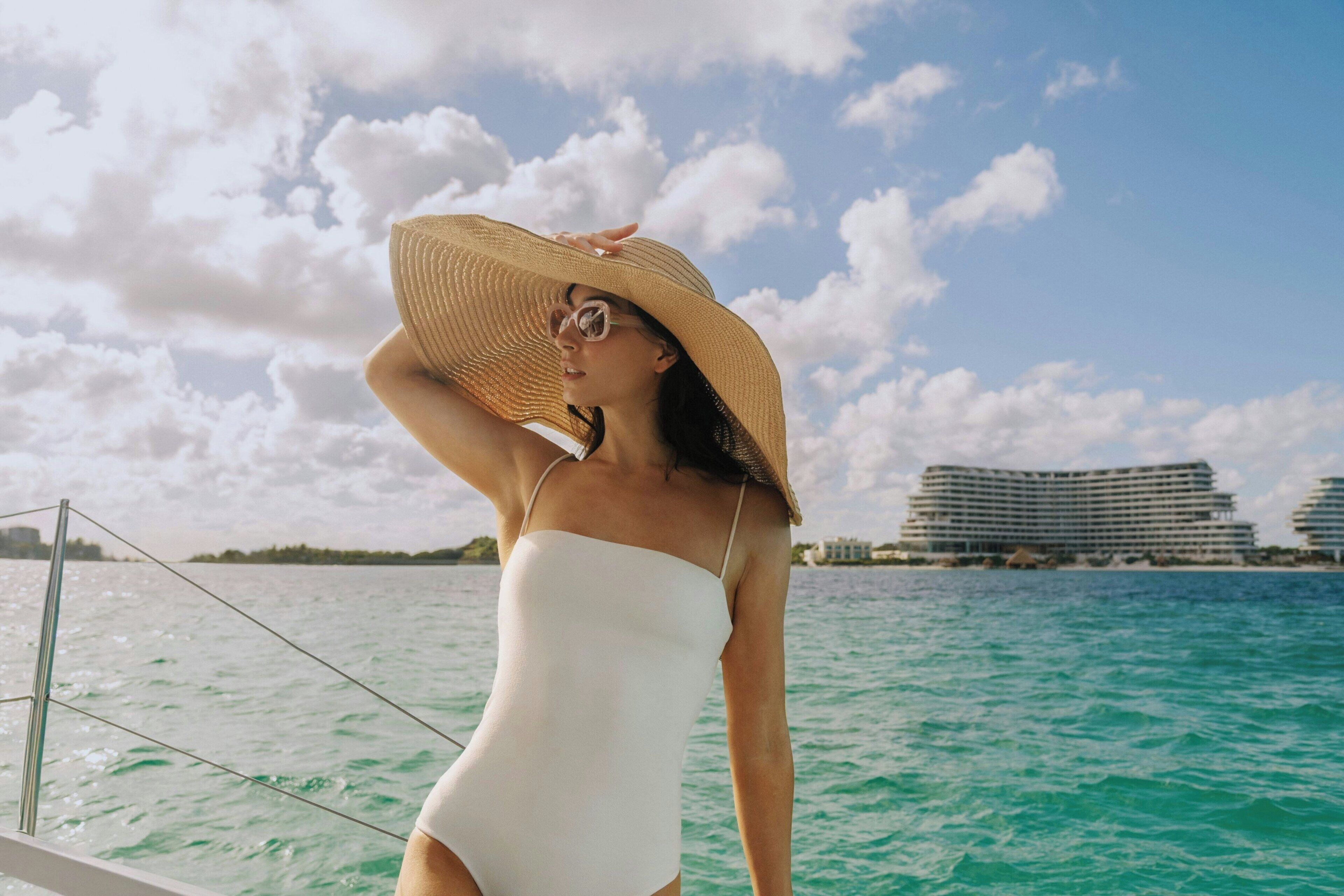 A person in a white swimsuit and straw hat on a boat with a view of a hotel and water.
