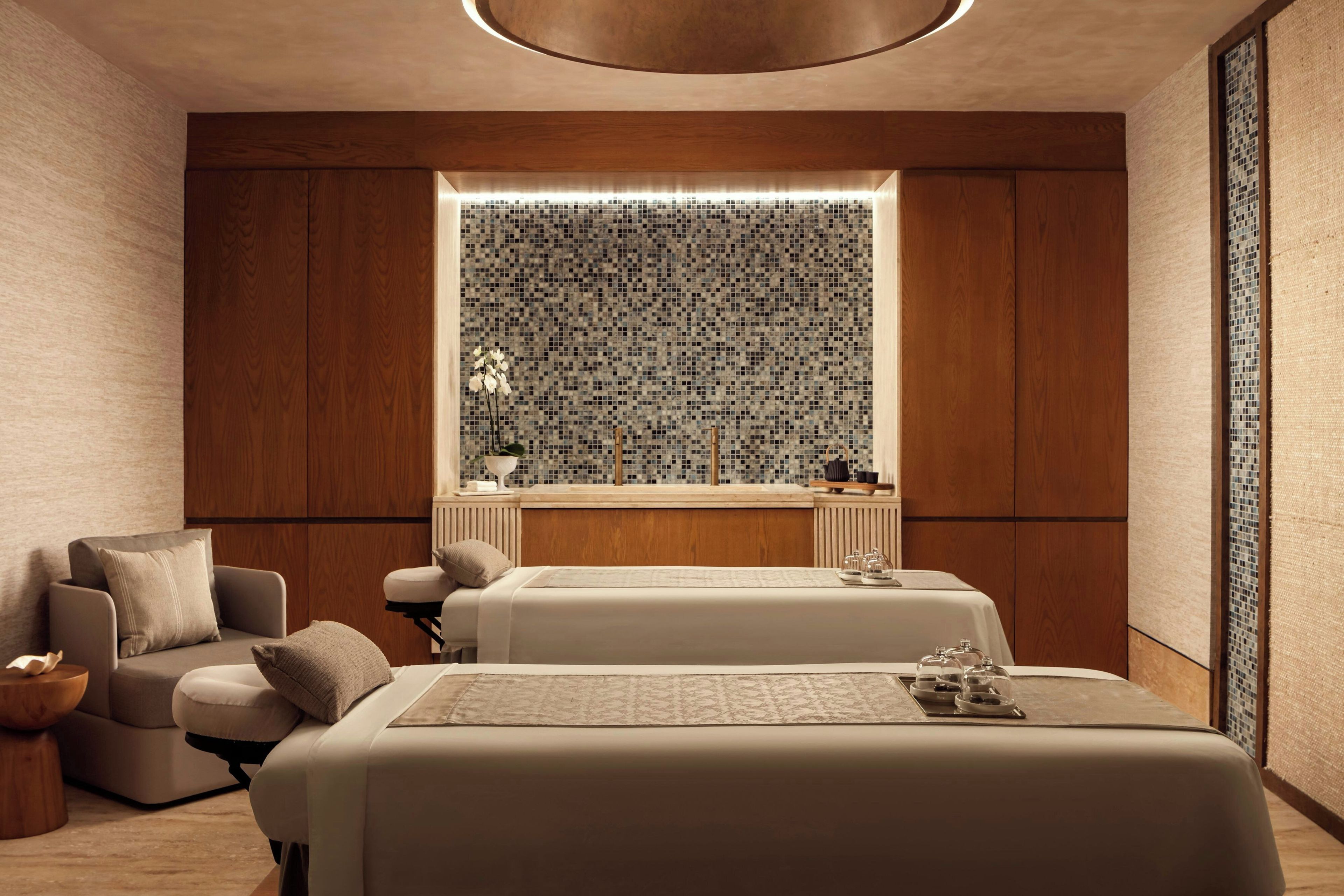 Un moderno salón de spa con dos camillas de masaje, un rincón de descanso con una silla, y un ventanal decorativo con un mosaico en la parte posterior.