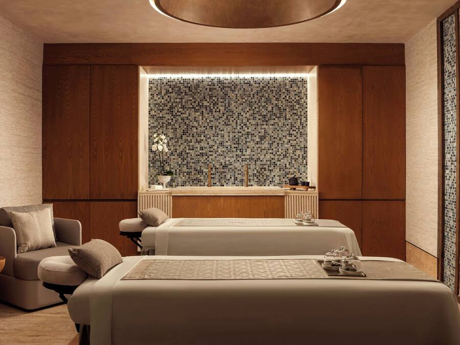 Un moderno salón de spa con dos camillas de masaje, un rincón de descanso con una silla, y un ventanal decorativo con un mosaico en la parte posterior.
