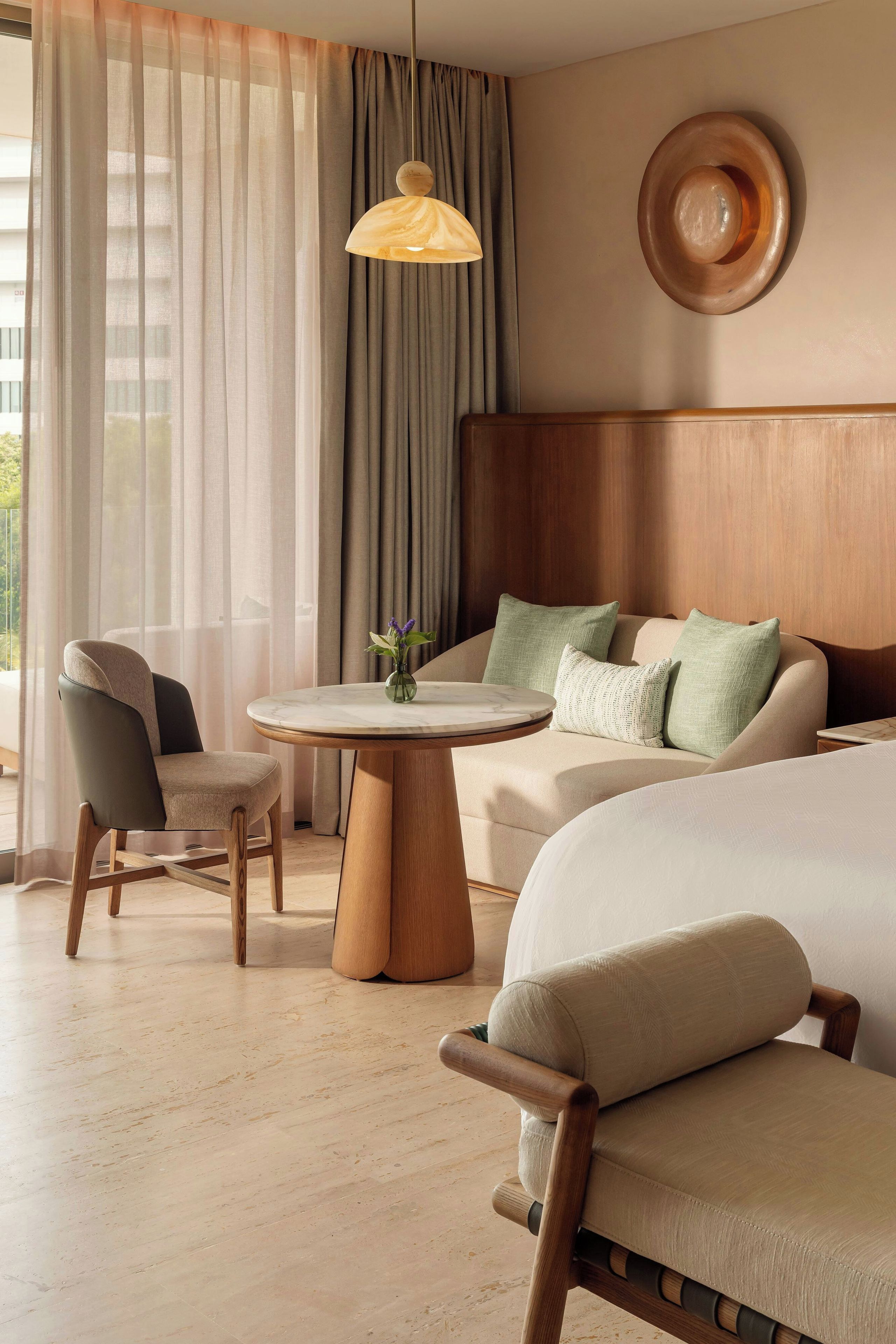 Habitación de hotel con cama, sofá, sillón, una mesa redonda de madera y una lámpara colgante.