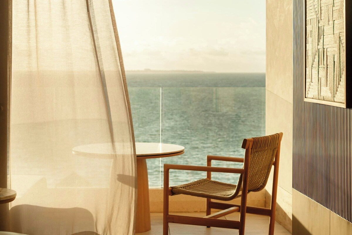 A room with a view of the sea, a chair, a table, and a bed.