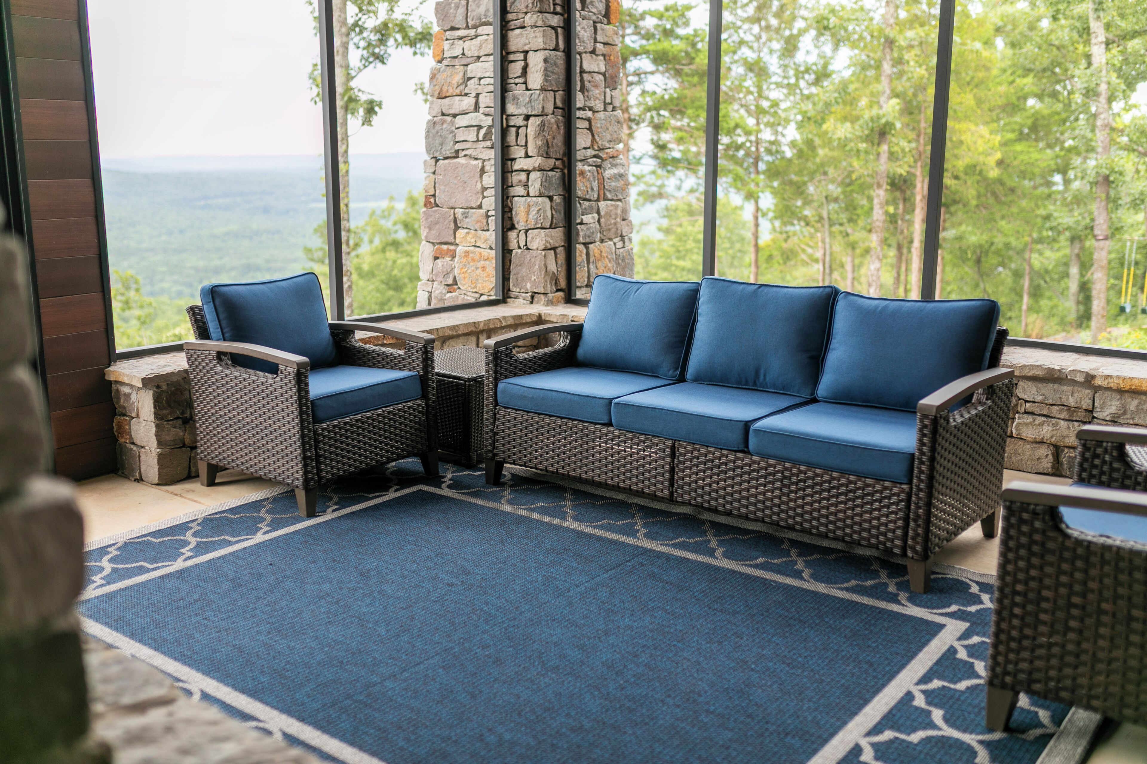 A wicker sofa with blue cushions, a matching armchair, and a small table in a room with a stone pillar and large windows overlooking a forested area.