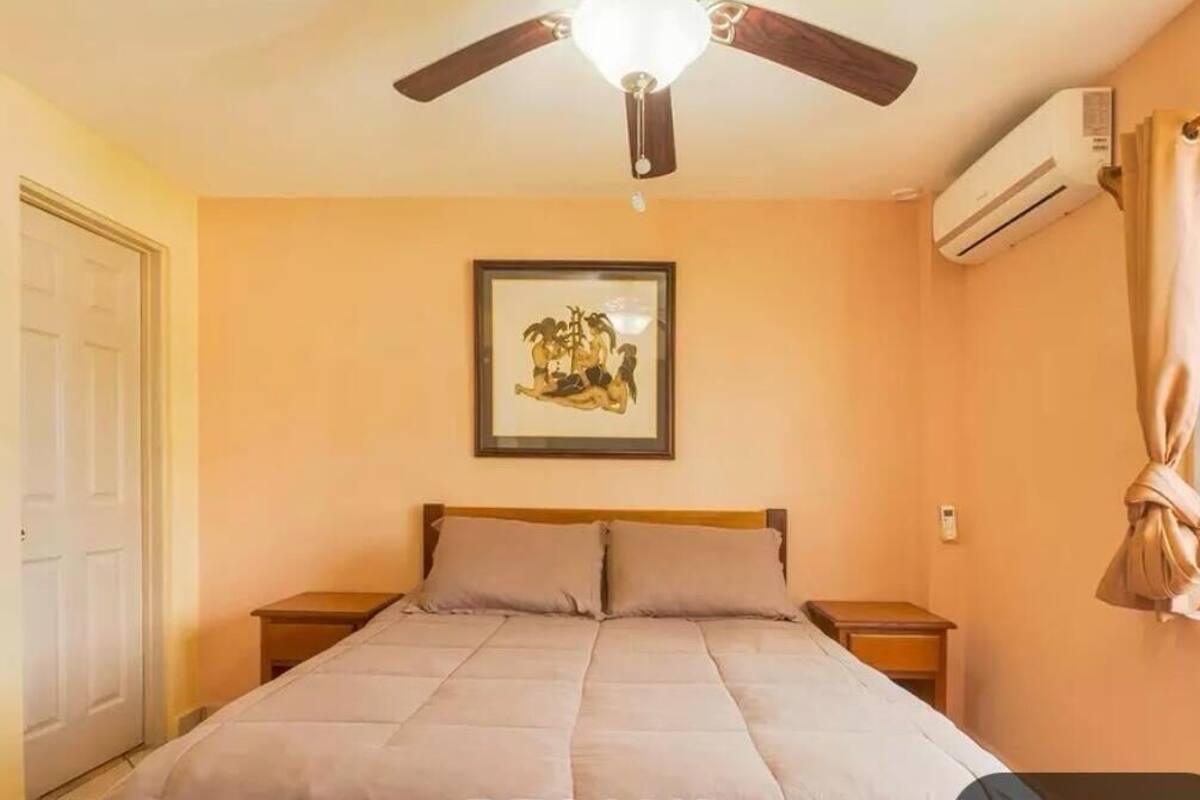 A bedroom with a bed, a ceiling fan, an air conditioning unit, a framed picture on the wall, and a window with curtains.
